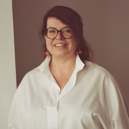 Eine Frau in einem weißen Hemd, mit Brille und roten Ohrringen, lächelt direkt in die Kamera.