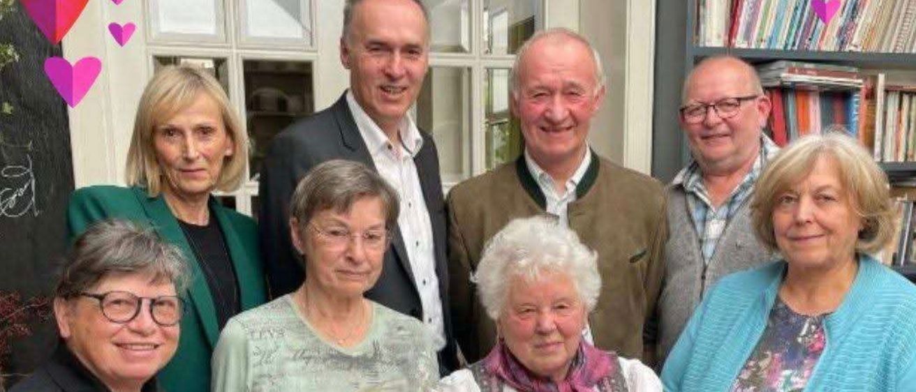 Eine Gruppe von sieben Personen posiert für ein Foto in einem Raum mit einem Bücherregal und Glasfenstern. Sie halten Blumen, und der Raum ist mit herzförmigen Motiven dekoriert.