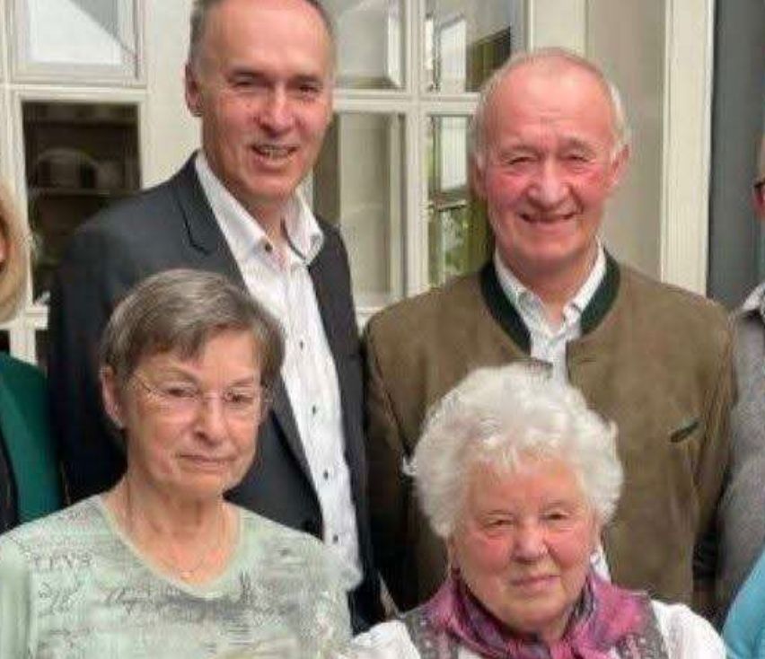 Eine Gruppe von sieben Personen posiert für ein Foto in einem Raum mit einem Bücherregal und Glasfenstern. Sie halten Blumen, und der Raum ist mit herzförmigen Motiven dekoriert.