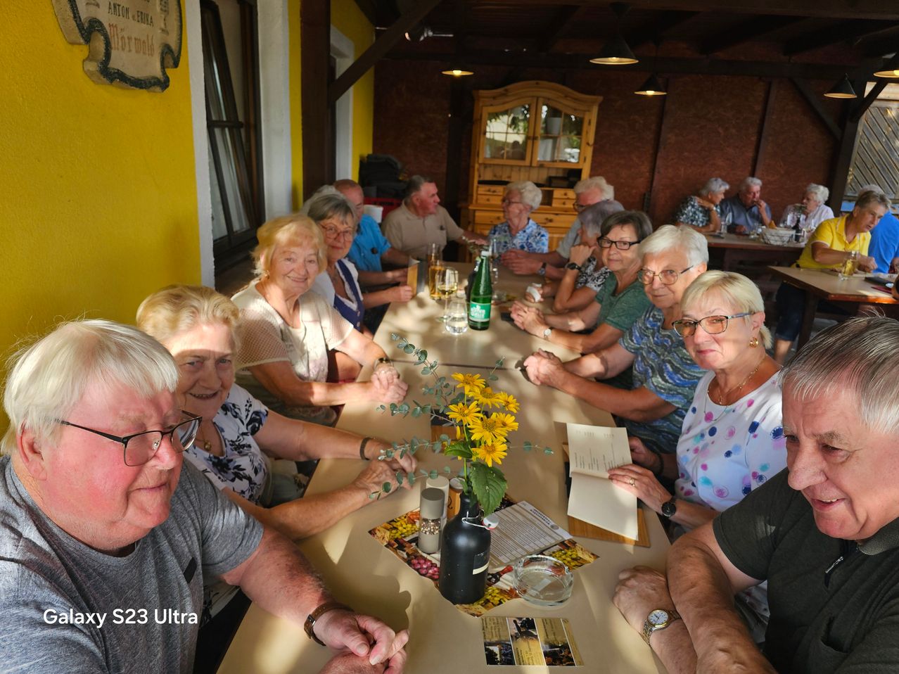 A group of elderly people sitting around a table in a restaurant, smiling and looking at the camera. There are flowers, a bottle, and a menu on the table.