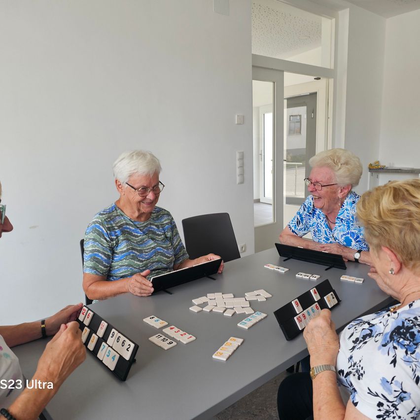 Four elderly women are sitting around a table, smiling and playing a game of mahjong. The table is filled with tiles and a black tray.