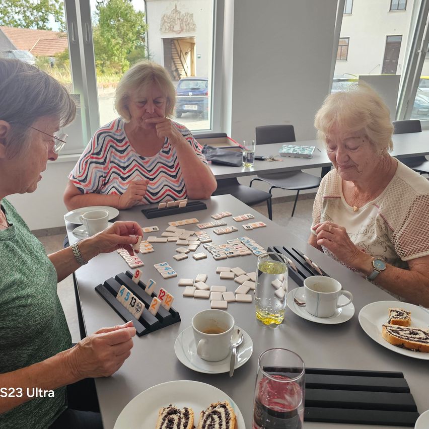 Three elderly women are sitting around a table, playing a game of tiles. One woman is looking at the tiles while another woman is smiling. There are cups, plates, and a bottle on the table.