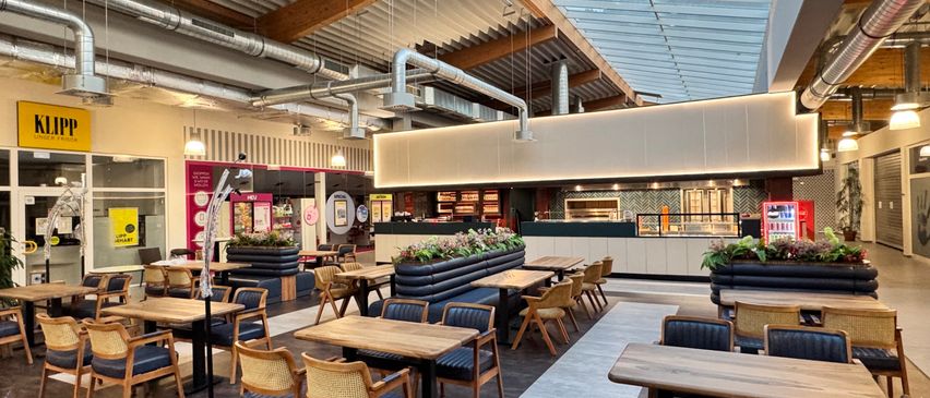 Ein Indoor-Restaurant mit Holztischen und Stühlen, einer Glasdecke und einer Theke mit einer Lebensmittelausstellung.