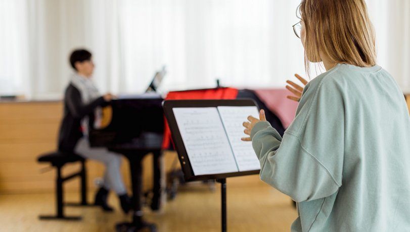 Eine Frau steht vor einem Notenständer mit Noten, während eine andere Person im Hintergrund Klavier spielt.