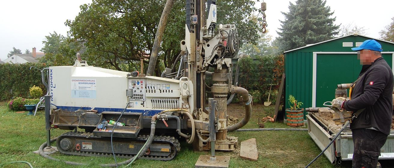 Eine Bohrmaschine steht auf dem Rasen, mit Bäumen im Hintergrund. Rechts befindet sich ein grüner Schuppen.