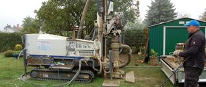 Eine Bohrmaschine steht auf dem Rasen, mit Bäumen im Hintergrund. Rechts befindet sich ein grüner Schuppen.