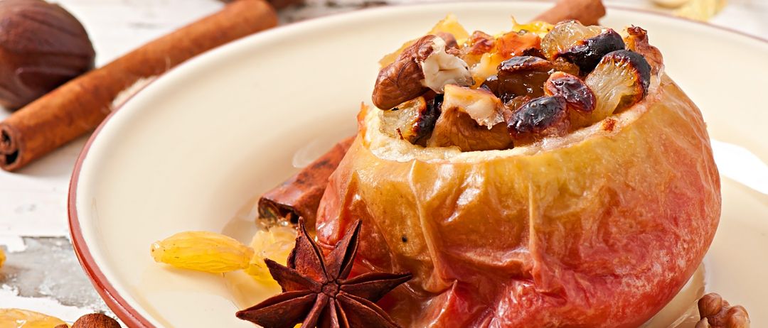 Ein festlicher Nachtisch auf einem weißen Teller, gefüllt mit einem roten Apfel, garniert mit Nüssen, Rosinen und einer Zimtstange. Im Hintergrund befinden sich goldene Dekorationen und eine Nuss.