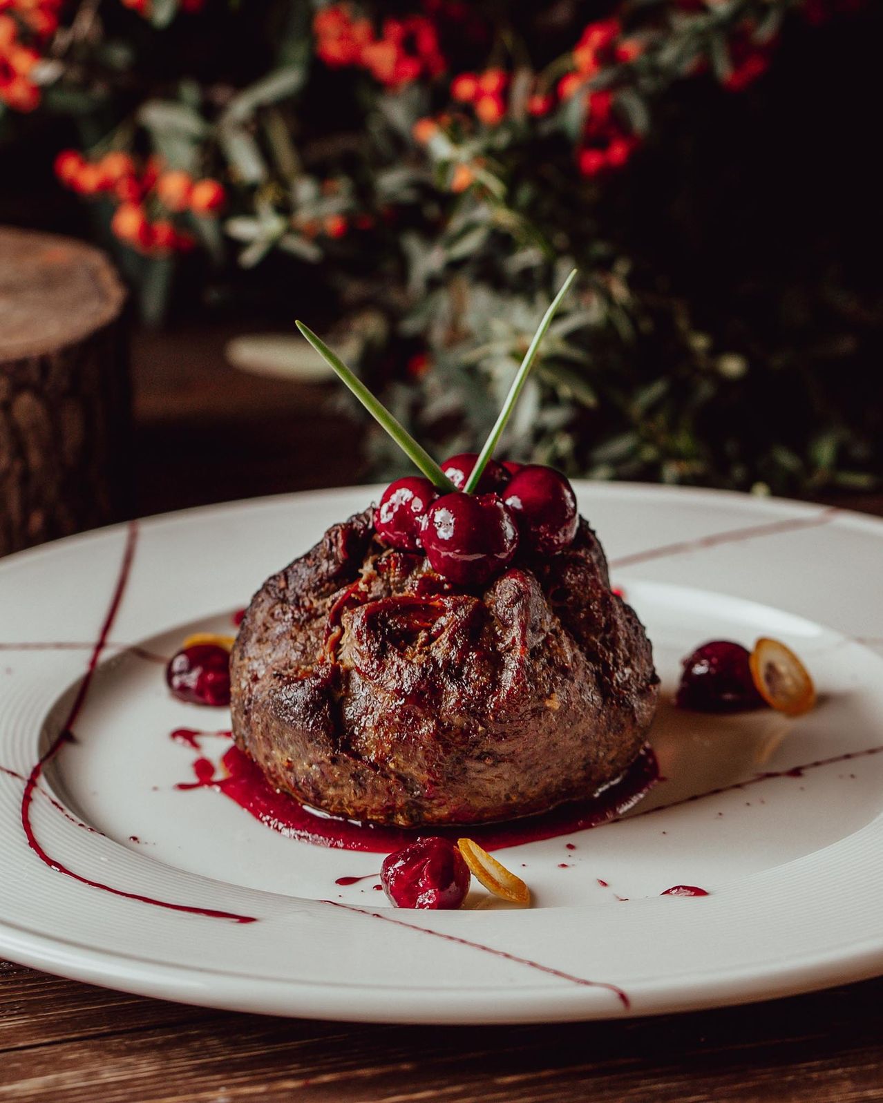Nahaufnahme eines Steakgerichts auf einem weißen Teller mit roter Soße und mit Kirschen und Kräutern garniert, mit einem verschwommenen Hintergrund roter Blumen.