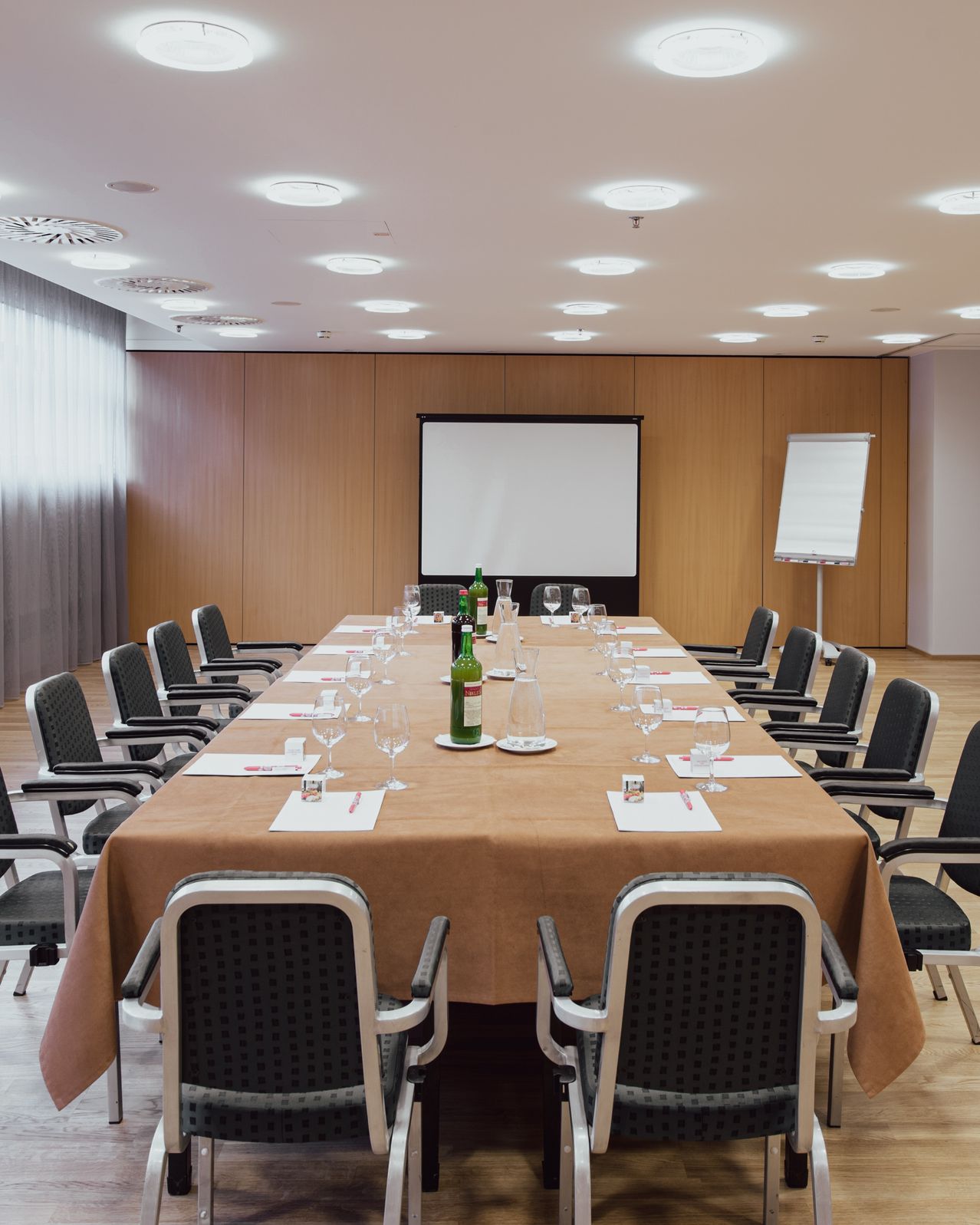 Ein leerer Konferenzraum mit einem langen Tisch, Stühlen, Weingläsern und zwei grünen Flaschen. Eine Whiteboard und eine Projektionsleinwand sind an der Wand.