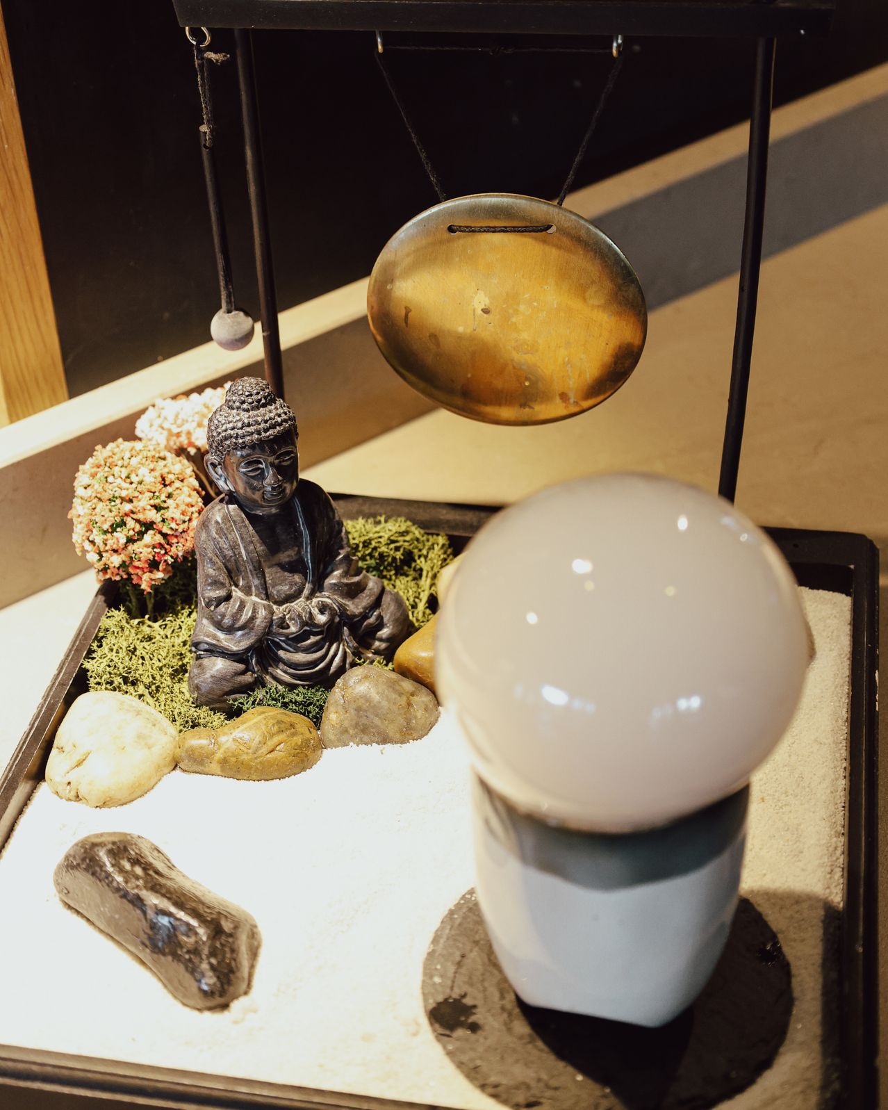 Eine Buddha-Statue sitzt in einer Schale mit Steinen, Moos, Blumen und einer hängenden Glocke. Eine weiße Kugel steht auf einem zylindrischen Sockel.