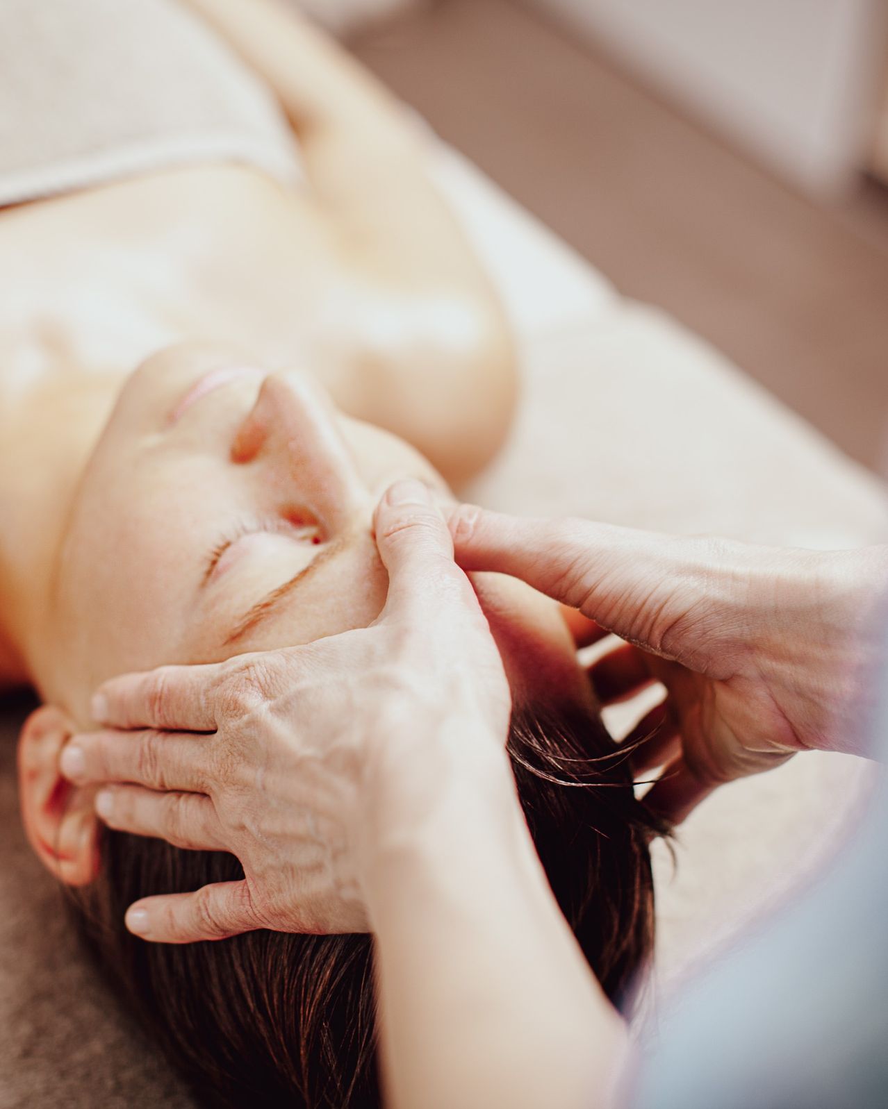 Eine Frau liegt auf einem Massagetisch und erhält von einem Therapeuten eine Gesichtsmassage. Die Hände des Therapeuten sind um das Gesicht der Frau positioniert und üben sanften Druck aus.