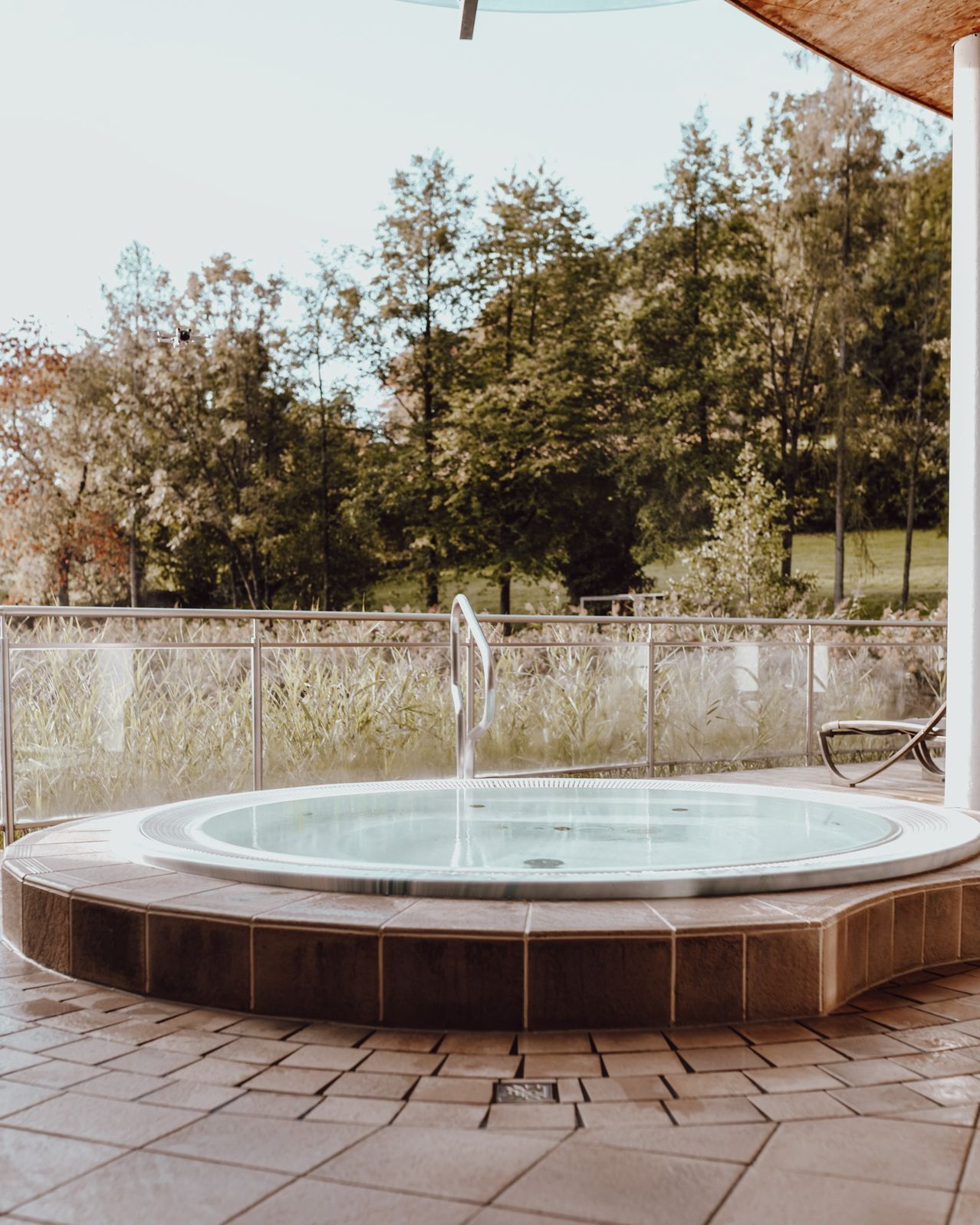 A circular hot tub on a brick patio, surrounded by a metal fence, with trees and a grassy field in the background.