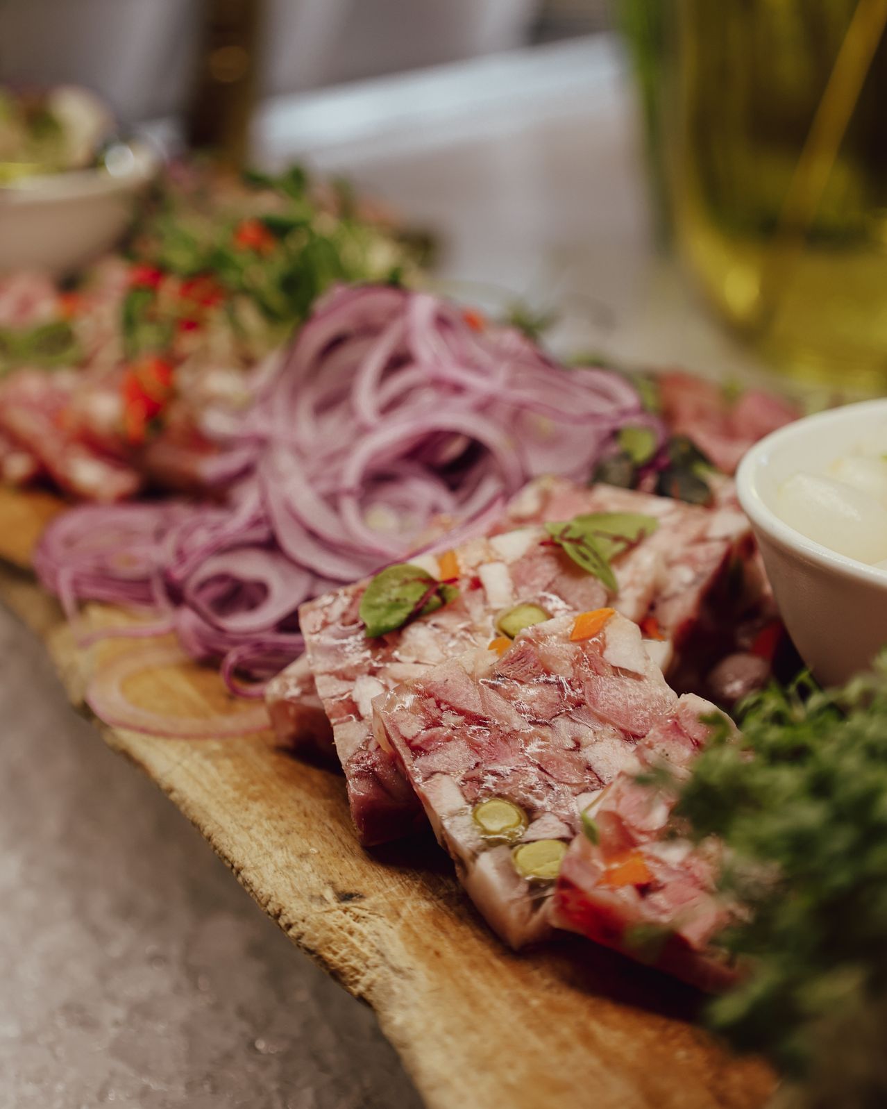 Ein Holzbrett zeigt Scheiben von gepökeltem Fleisch, garniert mit Kräutern und roten Zwiebeln, begleitet von einer Schüssel cremiger Dip.