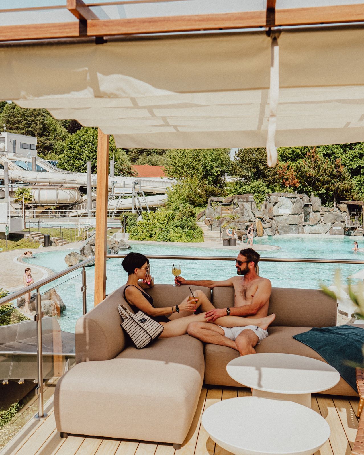 Zwei Personen sitzen auf einer Couch am Pool. Sie halten Getränke in den Händen und schauen in die Kamera. Im Hintergrund ist eine Wasserrutsche zu sehen.
