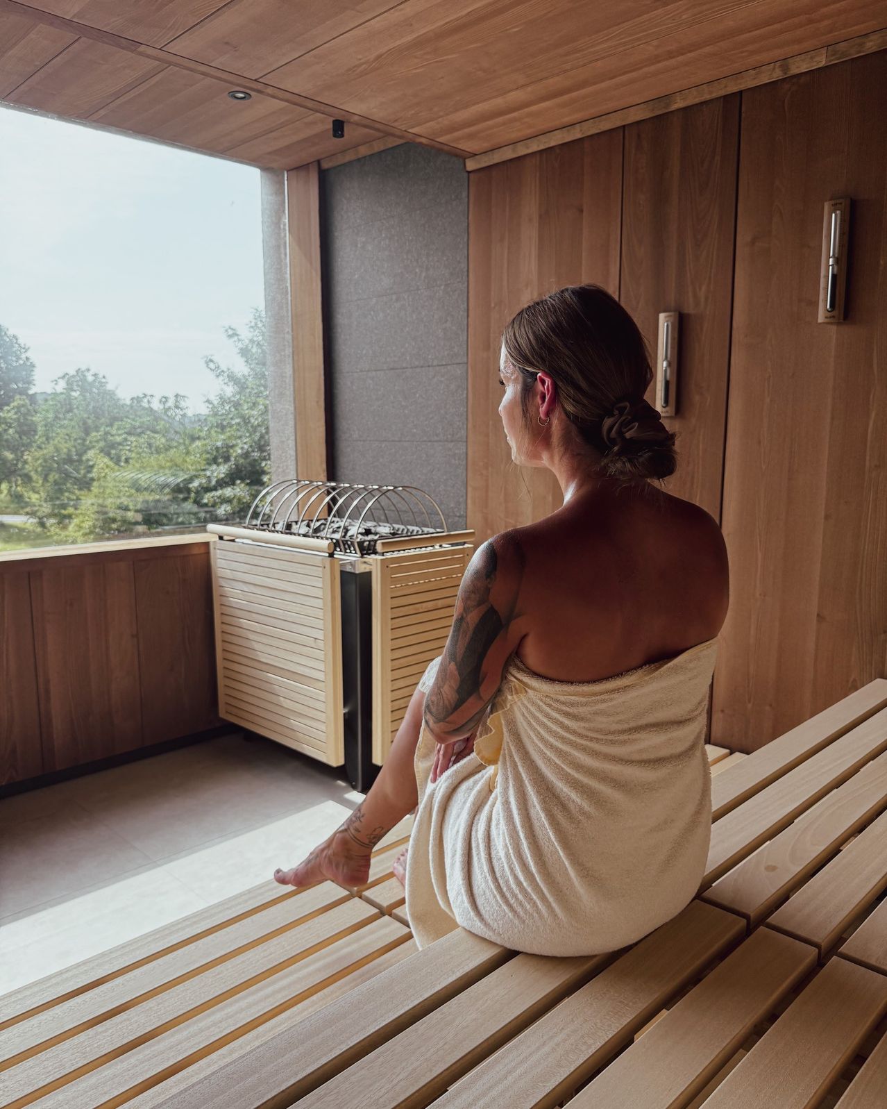 Eine Frau mit Tattoos sitzt in einer Holzsauna und schaut aus dem Fenster auf Bäume. Sie trägt ein weißes Handtuch und sitzt auf einer Bank.