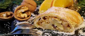 Ein festliches Dessert, bestehend aus einer Scheibe Apfelstrudel, bestreut mit Puderzucker, mit einer Gabel in der Nähe, und garniert mit Walnüssen und Orangenscheiben.