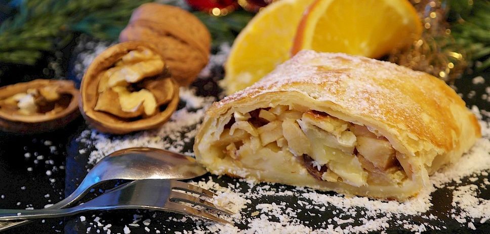 Ein festliches Dessert, bestehend aus einer Scheibe Apfelstrudel, bestreut mit Puderzucker, mit einer Gabel in der Nähe, und garniert mit Walnüssen und Orangenscheiben.