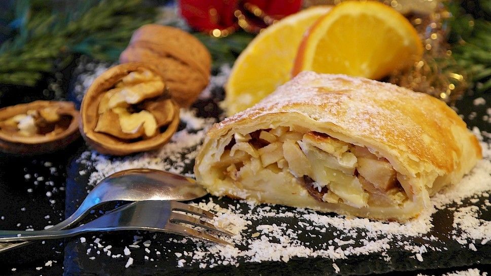 Ein festliches Dessert, bestehend aus einer Scheibe Apfelstrudel, bestreut mit Puderzucker, mit einer Gabel in der Nähe, und garniert mit Walnüssen und Orangenscheiben.