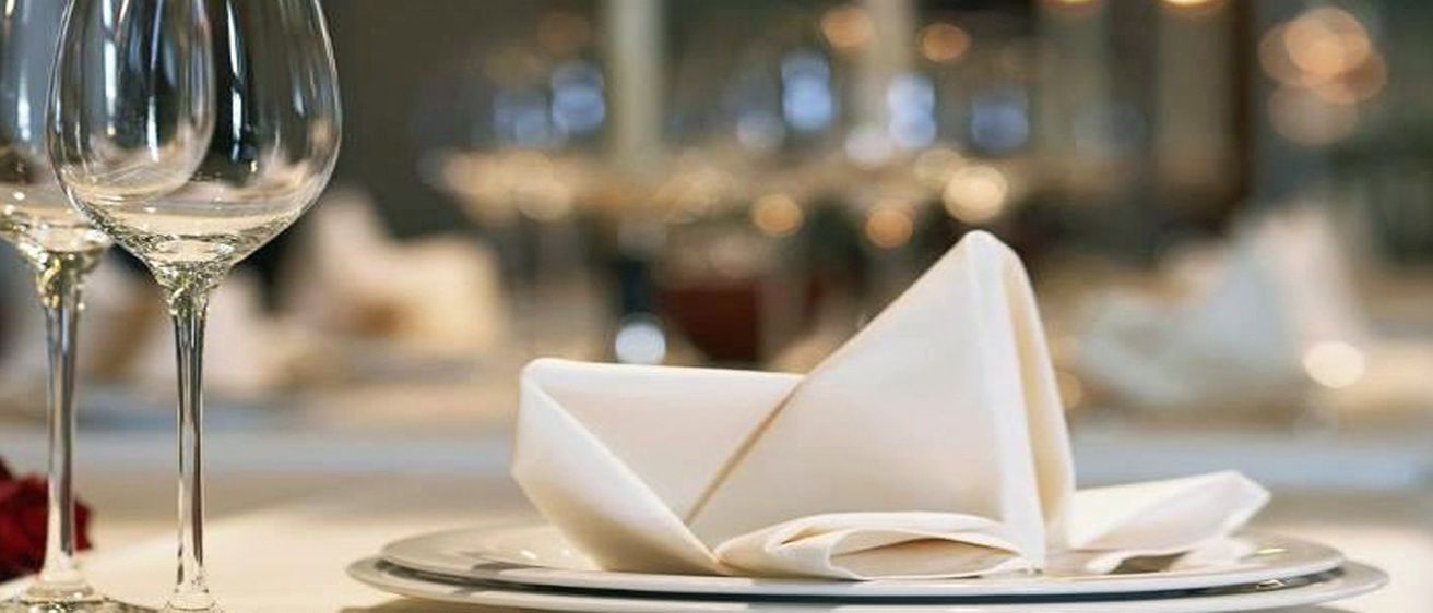 Ein ordentlich gefaltetes weißes Serviettentuch liegt auf einem Teller, mit verschwommenen Weingläsern und einem Tisch im Hintergrund, was auf ein formelles Ambiente hindeutet.