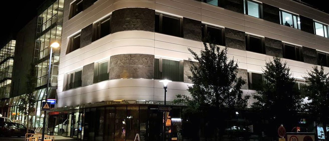 Ein modernes Gebäude mit strukturierten Steinakzenten, gut beleuchtet in der Nacht. Eine Straßenlampe und ein Baum stehen davor.