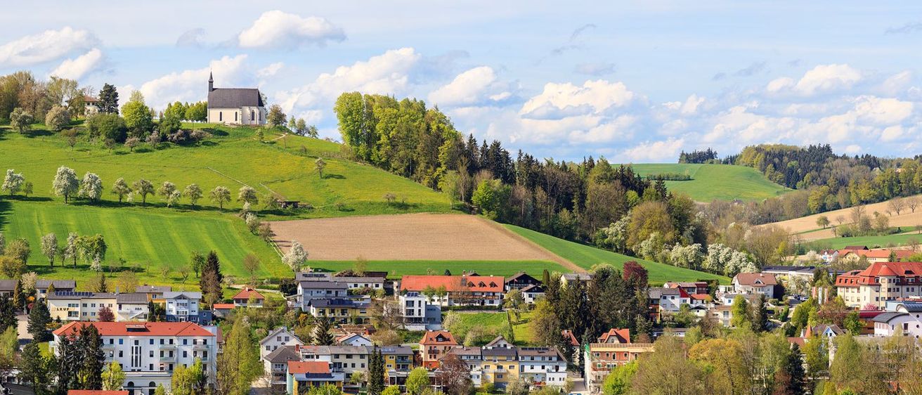 Ein Dorf mit farbenfrohen Häusern, eingebettet in einem Tal, umgeben von üppigen grünen Feldern und Bäumen. Eine Kirche steht auf einem Hügel über dem Dorf.