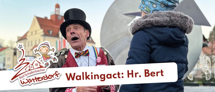 Ein Mann in einem bunten Kostüm mit einem Zylinder spricht mit einem Kind, das eine blaue Jacke und eine Kopfbedeckung trägt. Der Schriftzug auf dem Banner lautet 'Walking Act: Mr. Bert'.