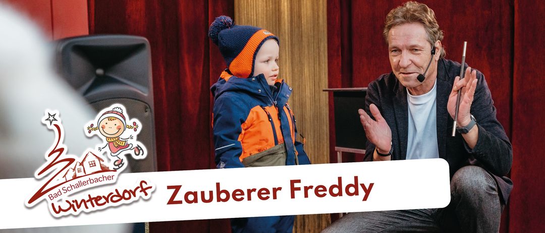 Ein Mann mit Mikrofon sitzt neben einem Kind in einer orange-blauen Jacke. Hinter ihnen ist ein großer Lautsprecher mit einem Cartoon-Aufkleber zu sehen, und sie stehen vor einem roten Vorhang.