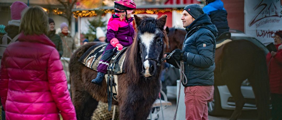 Ein kleines Kind reitet ein Pferd in einem Park. Ein Mann hält die Zügel, während eine Frau und andere zusehen. Der Hintergrund ist festlich mit Dekorationen und einem Bauernhof.