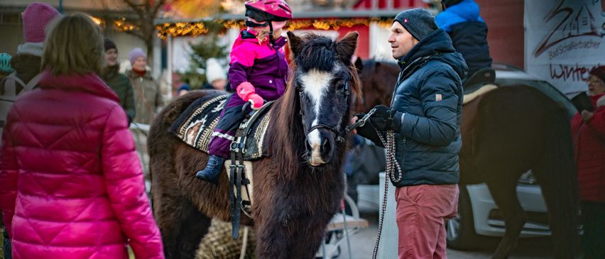 Ein kleines Kind reitet ein Pferd in einem Park. Ein Mann hält die Zügel, während eine Frau und andere zusehen. Der Hintergrund ist festlich mit Dekorationen und einem Bauernhof.