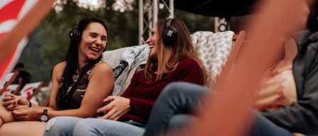 Zwei Frauen mit Kopfhörern und lächelnd, sitzen auf einer Bank bei einer Veranstaltung im Freien. Sie scheinen Musik zu genießen. Bäume und eine Bühne mit einem Vordach sind im Hintergrund.