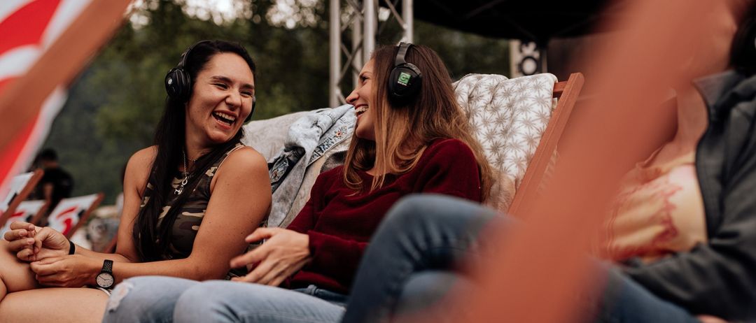 Zwei Frauen mit Kopfhörern und lächelnd, sitzen auf einer Bank bei einer Veranstaltung im Freien. Sie scheinen Musik zu genießen. Bäume und eine Bühne mit einem Vordach sind im Hintergrund.