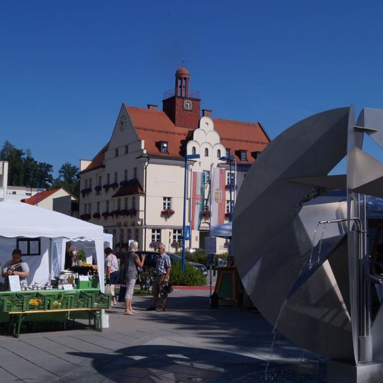 Ein Marktplatz mit Ständen unter einem weißen Zelt, bei dem Menschen stehen und reden. Ein moderner Brunnen fließt auf der Straße. Dahinter steht ein großes Gebäude mit einem Turm.