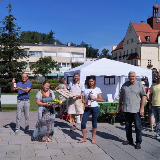Eine Gruppe von Menschen steht vor einem weißen Zelt. Eine Frau hält einen Bund Gemüse und eine Flasche, während eine andere Frau einen Teller mit Essen hält. Dahinter befindet sich ein Gebäude mit einem Glockenturm.
