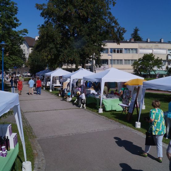 Ein Outdoor-Marktereignis in einem Park mit verschiedenen Zelten, Käufern und Verkäufern. Bäume und Gebäude sind im Hintergrund sichtbar.