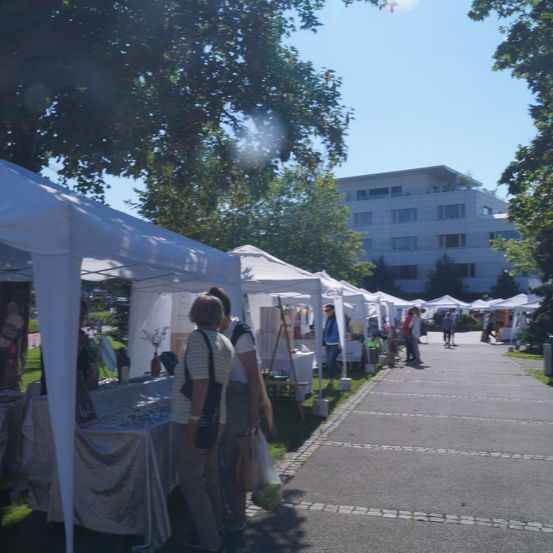 Ein Freiluftmarkt mit weißen Zelten an einem sonnigen Tag. Mehrere Menschen laufen und stöbern. Ein Gebäude mit vielen Fenstern ist im Hintergrund.