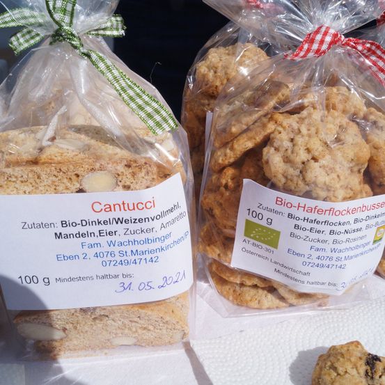 Zwei Plastiktüten mit grünen und roten Bändern enthalten Kekse. Eine Tüte ist mit 'Cantucci' beschriftet und enthält Zutaten wie Roggenmehl und Mandeln, mit einem Mindesthaltbarkeitsdatum vom 31. Mai 2021. Die andere Tüte ist mit 'Bio-Haferflockenbüssel' beschriftet und enthält Zutaten wie Haferflocken und Eier, mit einem Mindesthaltbarkeitsdatum vom 31. Mai 2021.