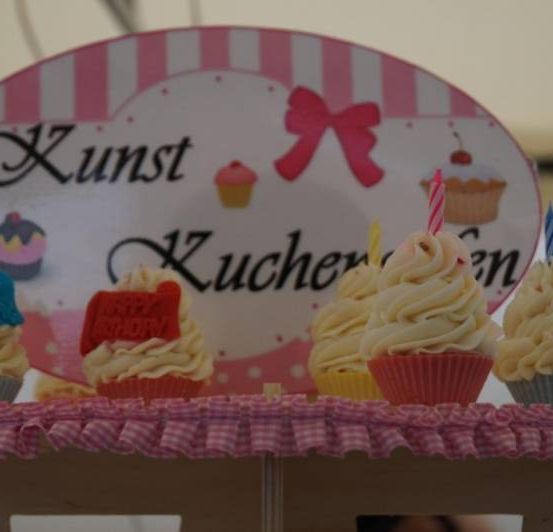 Fünf Cupcakes mit gelber Glasur und Kerzen stehen auf einem Tisch mit einem pink-weißen Schild, auf dem Kunst Kuchen steht. Es gibt ein Cupcake-Bild mit einer Kirsche oben und einer Schleife auf dem Schild.