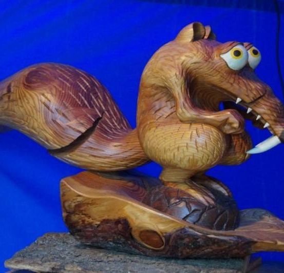 Eine hölzerne Eichhörnchenskulptur mit großen Augen und scharfen Zähnen sitzt auf einem Felsen vor einem blauen Hintergrund.