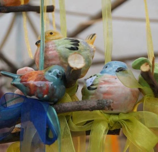 Mehrere farbenfrohe Keramikvögel hängen an einem Ast, jeder mit einem Band befestigt. Die Vögel haben unterschiedliche Farben und Designs.