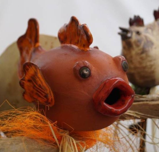 Ein Nahaufnahme einer roten Fischskulptur mit offenem Mund, umgeben von anderen Holzskulpturen, möglicherweise auf einem weißen Hintergrund ausgestellt.