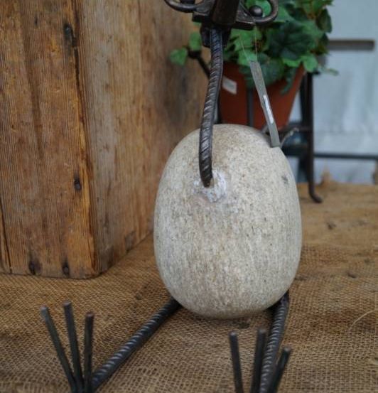 Ein dekoratives Stein-Ei mit Metallbeinen auf einer Holzoberfläche, mit einem Topfpflanzen im Hintergrund.