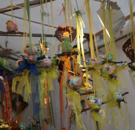Eine Sammlung bunter Vogelfiguren, die von der Decke hängen, jeweils auf einem Stock mit gelben Bändern. Ein herzförmiges Ornament mit einer Nachricht hängt ebenfalls.