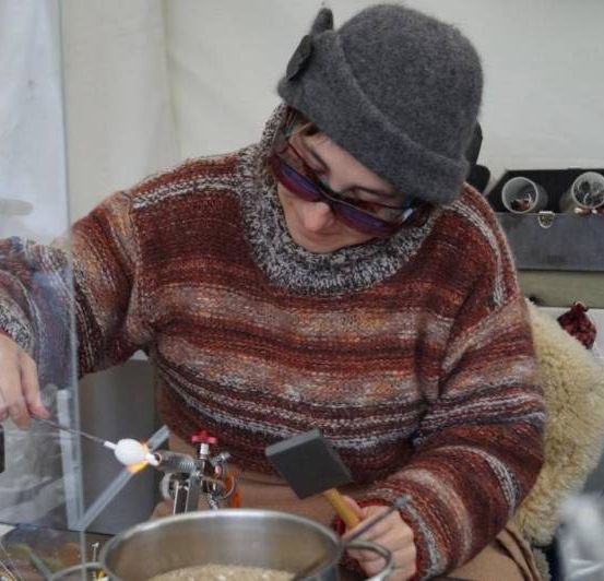 Eine Frau mit grauem Beanie und Brille arbeitet an einem Tisch, hält einen Hammer und einen Topf mit etwas darin. Sie benutzt einen Glasstab und eine Flamme. Dahinter ist eine weiße Wand und ein Schreibtisch mit verschiedenen Gegenständen.