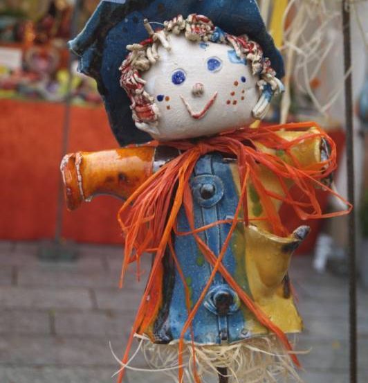 Ein bemalter Vogelscheuche steht auf einem Outdoor-Markt mit einem blauen Hut, gelber Jacke und orangefarbenen Bändern. Im Hintergrund ist ein roter Stoff zu sehen, und einige Bäume sind sichtbar.