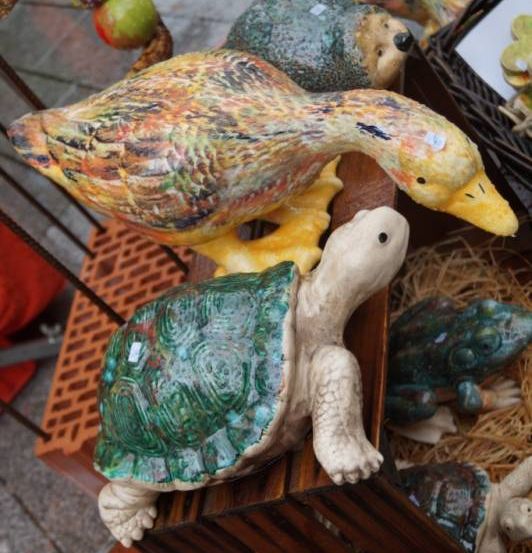 Zwei bemalte Keramikenten sitzen auf einer Holzkiste, darunter eine Schildkrötenfigur und weitere Figuren im Hintergrund.