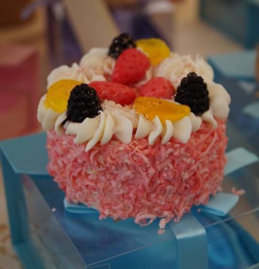 Ein kleiner rosa Kuchen mit weißer Glasur und einer Mischung aus Beeren und Zitrusfrüchten darauf ist in einer blauen Schachtel platziert.