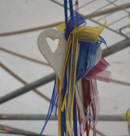 Ein weißes herzförmiges Objekt mit Bändern, die daran hängen, aufgehängt an einer Metallstruktur. Das Herz hat einen gelben Umriss und ist von mehrfarbigen Bändern umgeben.
