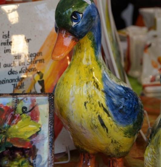 Eine farbenfrohe Keramik-Entenfigur steht auf einem Tisch neben einem gerahmten Kunstwerk und anderen Keramikartikeln. Die Ente hat einen gelben Körper, blaue Flügel und einen grünen Kopf. Der Hintergrund ist unscharf.