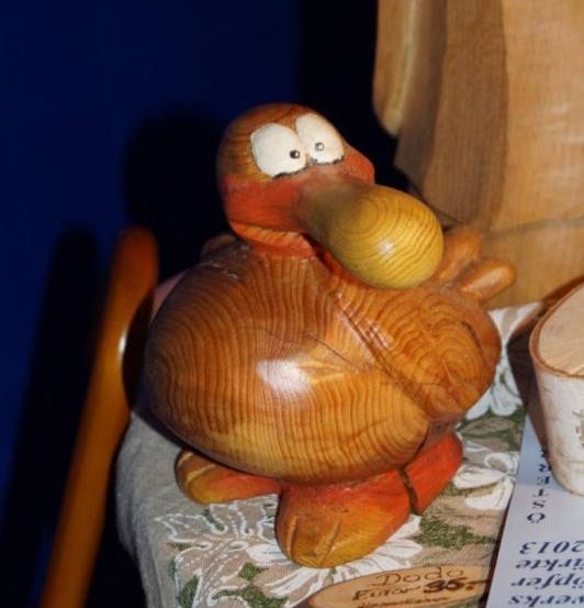 Eine hölzerne Entenfigur steht auf einem Tisch mit einer blumigen Tischdecke. Daneben befindet sich ein hölzerner Gehstock und ein blauer Zettel mit Text und Zahlen. Im Hintergrund ist ein teilweise sichtbares hölzerne Objekt.