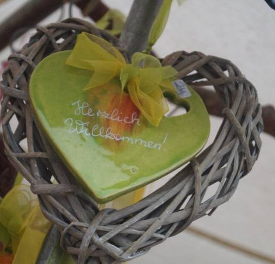 Ein grünes herzförmiges Keramikornament mit einem gelben Band, das Herzlich Willkommen! steht, hängt an einem Baum.